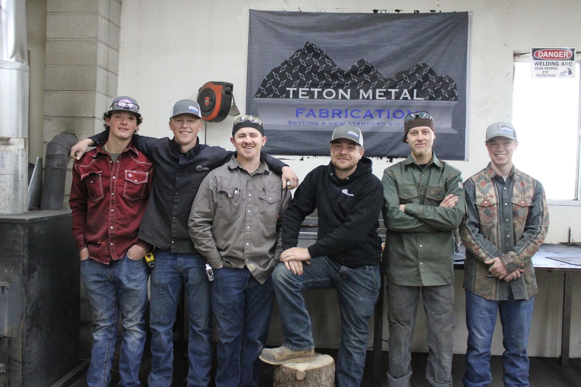 Six people pose in a metal fabrication shop, standing in front of a banner that reads