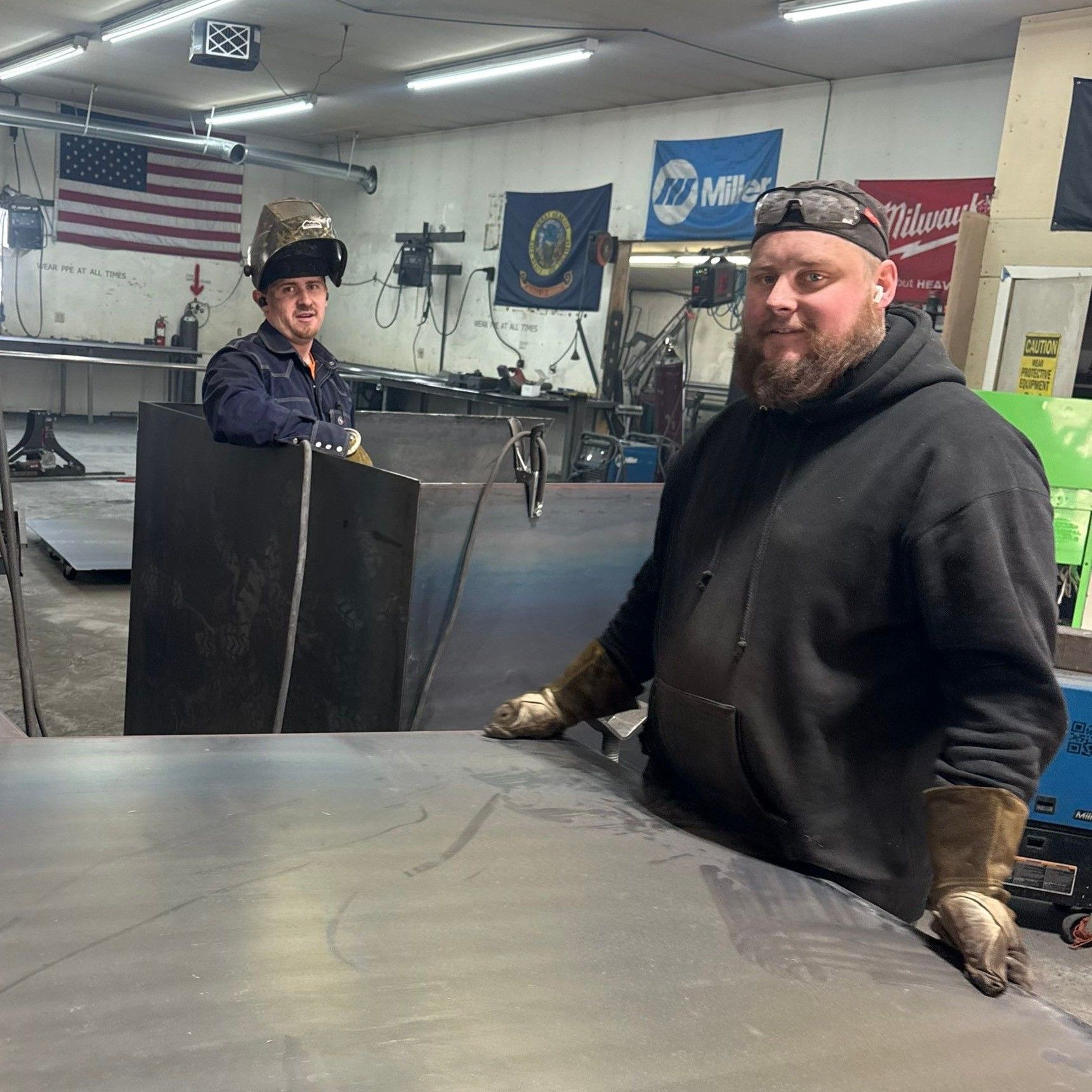 Two people in a metal workshop stand behind a large steel sheet; one wears a welding helmet, the other wears gloves.