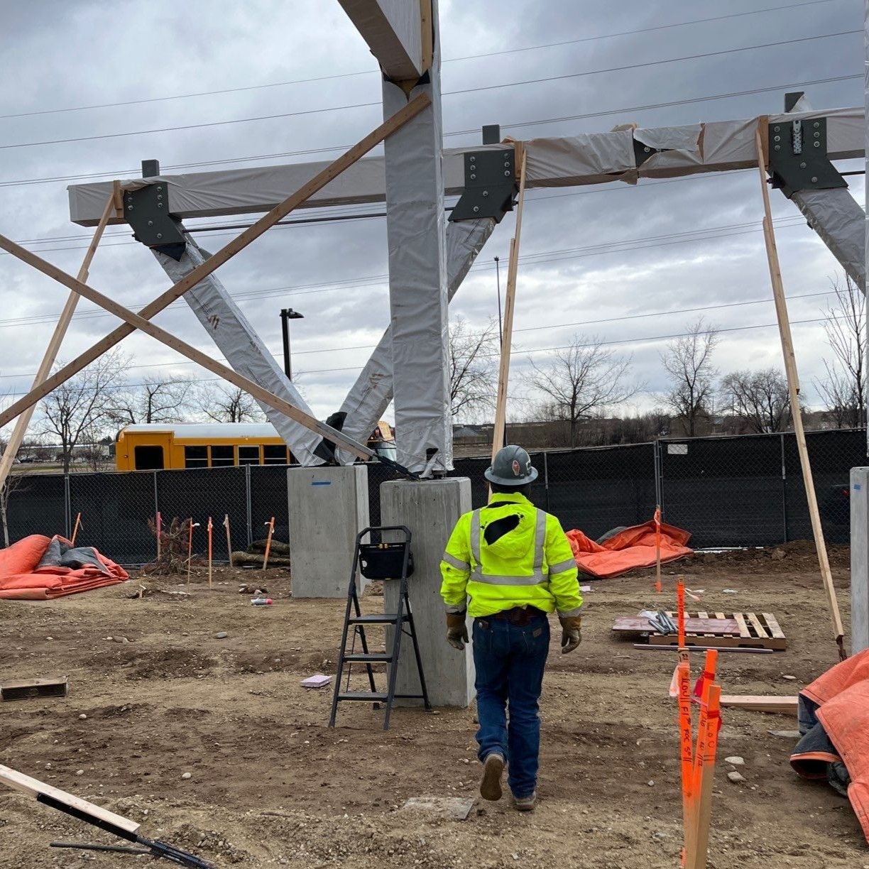 A construction worker in a high-visibility yellow vest walks toward a framework of partially installed wooden beams.