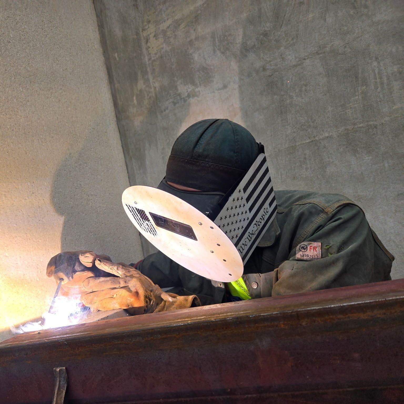 A welder wears a protective hood with a flag pattern while using a torch to weld a metal beam against a concrete wall.