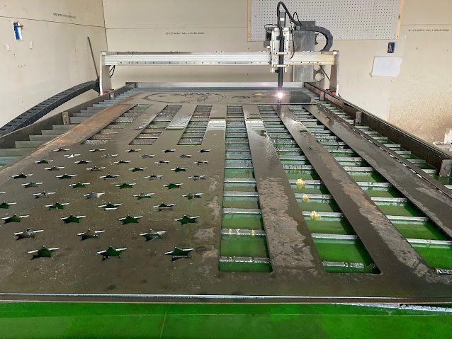 Metal sheet being cut by a plasma cutter, forming an American flag pattern. Green bed, indoor setting.