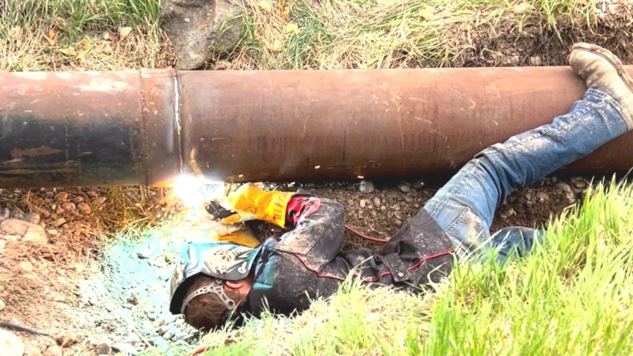 Person welding a large metal pipe in a trench, sparks flying.