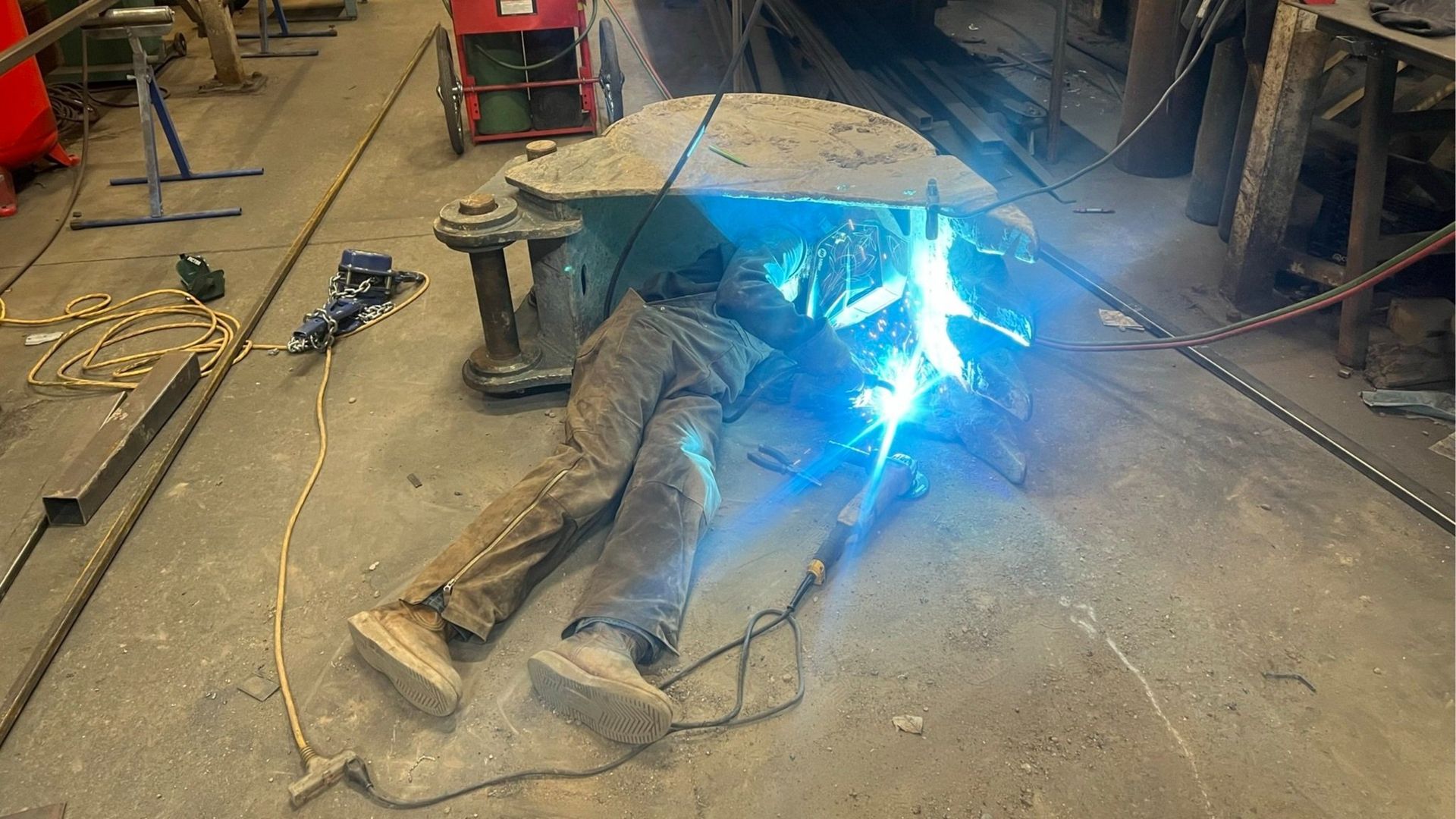 Welder lying on floor, welding a metal structure, sparks flying.