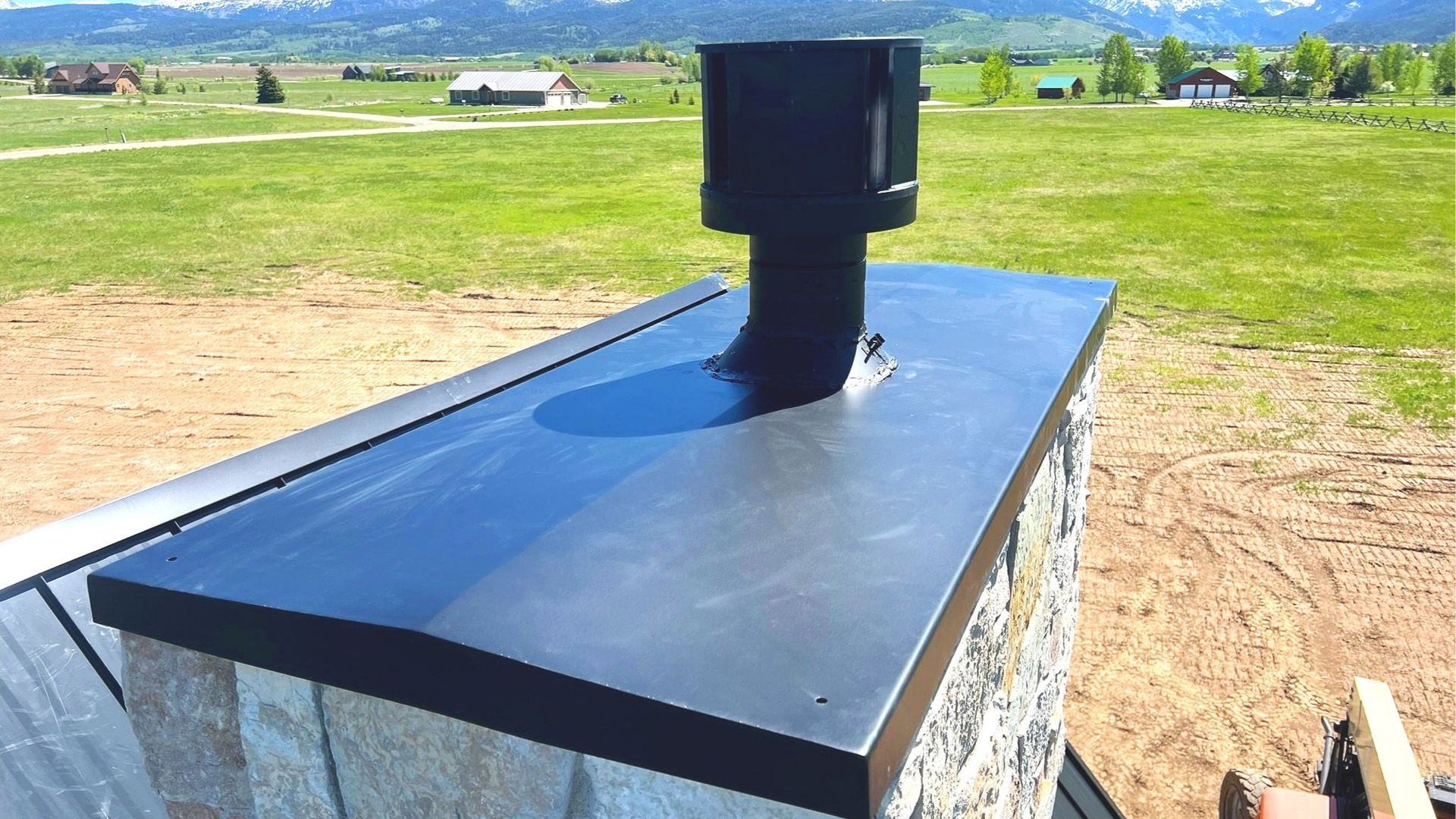 Black chimney vent on a stone chimney. Flat black metal roof cap with a grassy field and mountains in the background.