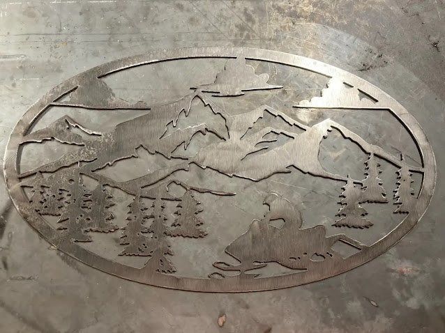 Metal cutout of mountains, trees, and a snowmobile inside an oval frame.