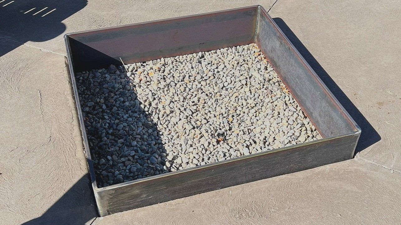Square metal container filled with gray gravel, on a concrete surface. Shadow on the left.