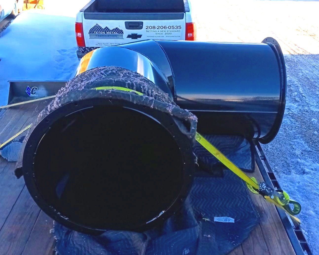 Black pipe elbow on a flatbed trailer, secured with a yellow strap, with a white truck in the background.