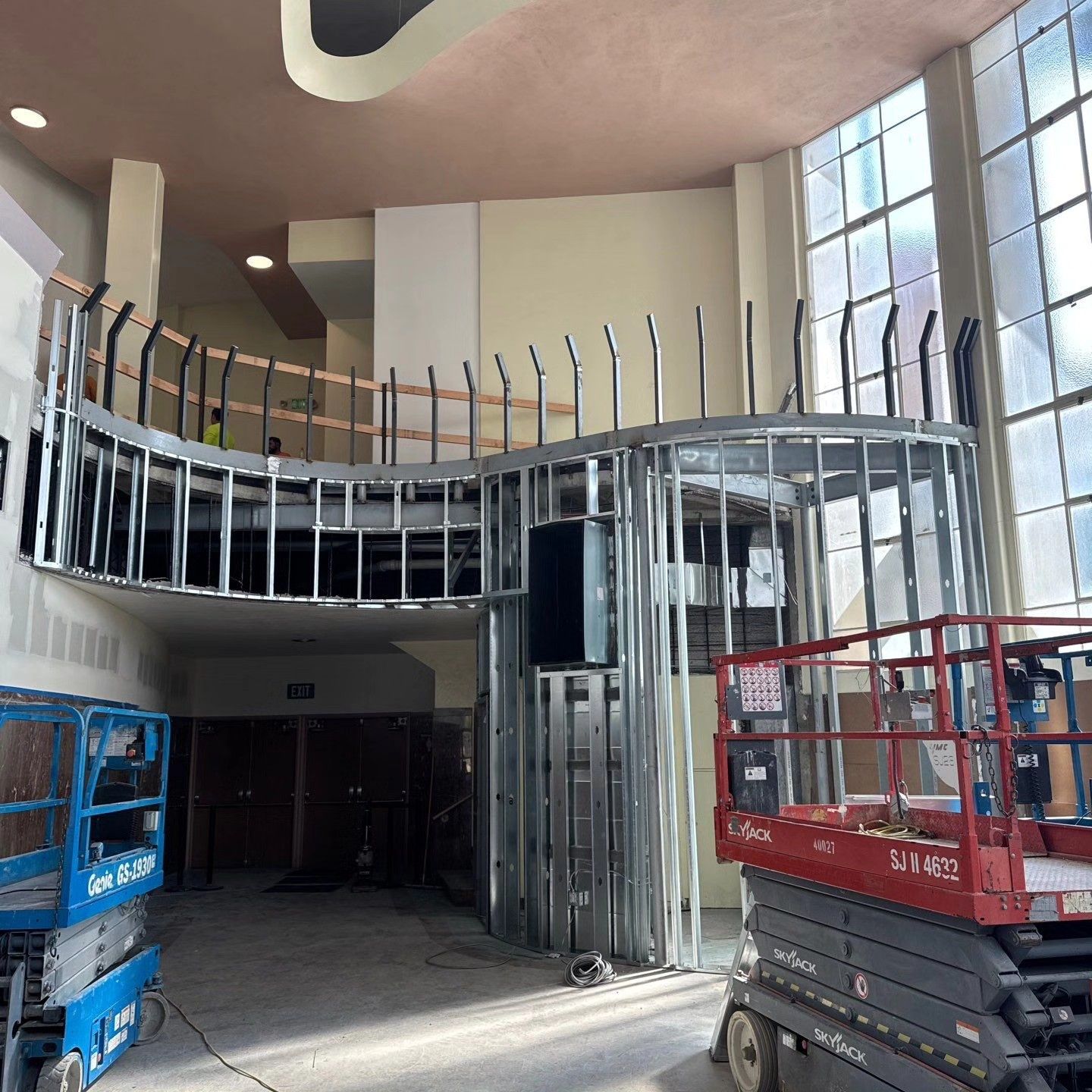 Interior construction: Metal framing for a curved balcony and support structure in a large room with windows and lift equipment.