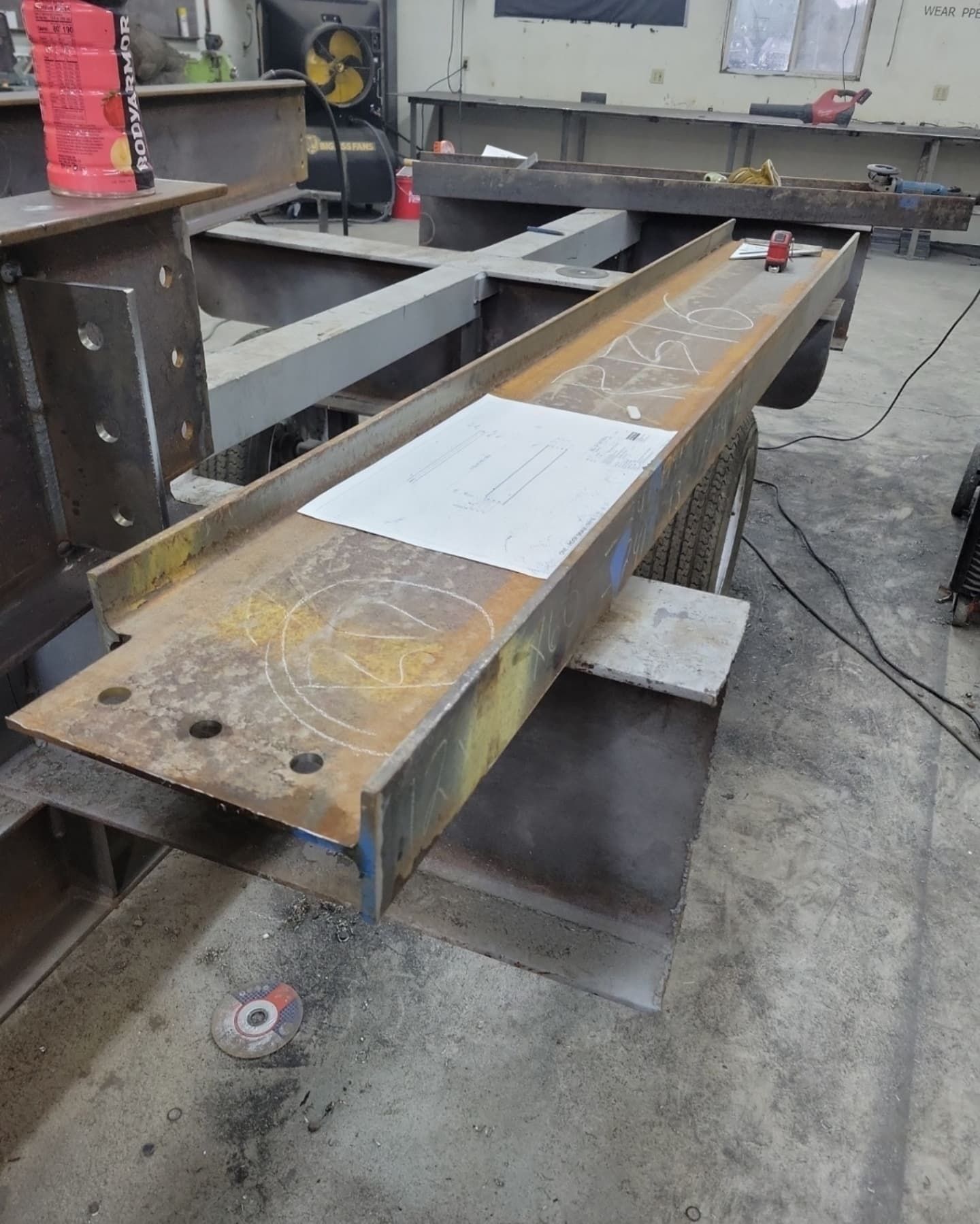A metal fabrication project with a long steel beam on a workbench. A blueprint rests on the beam.