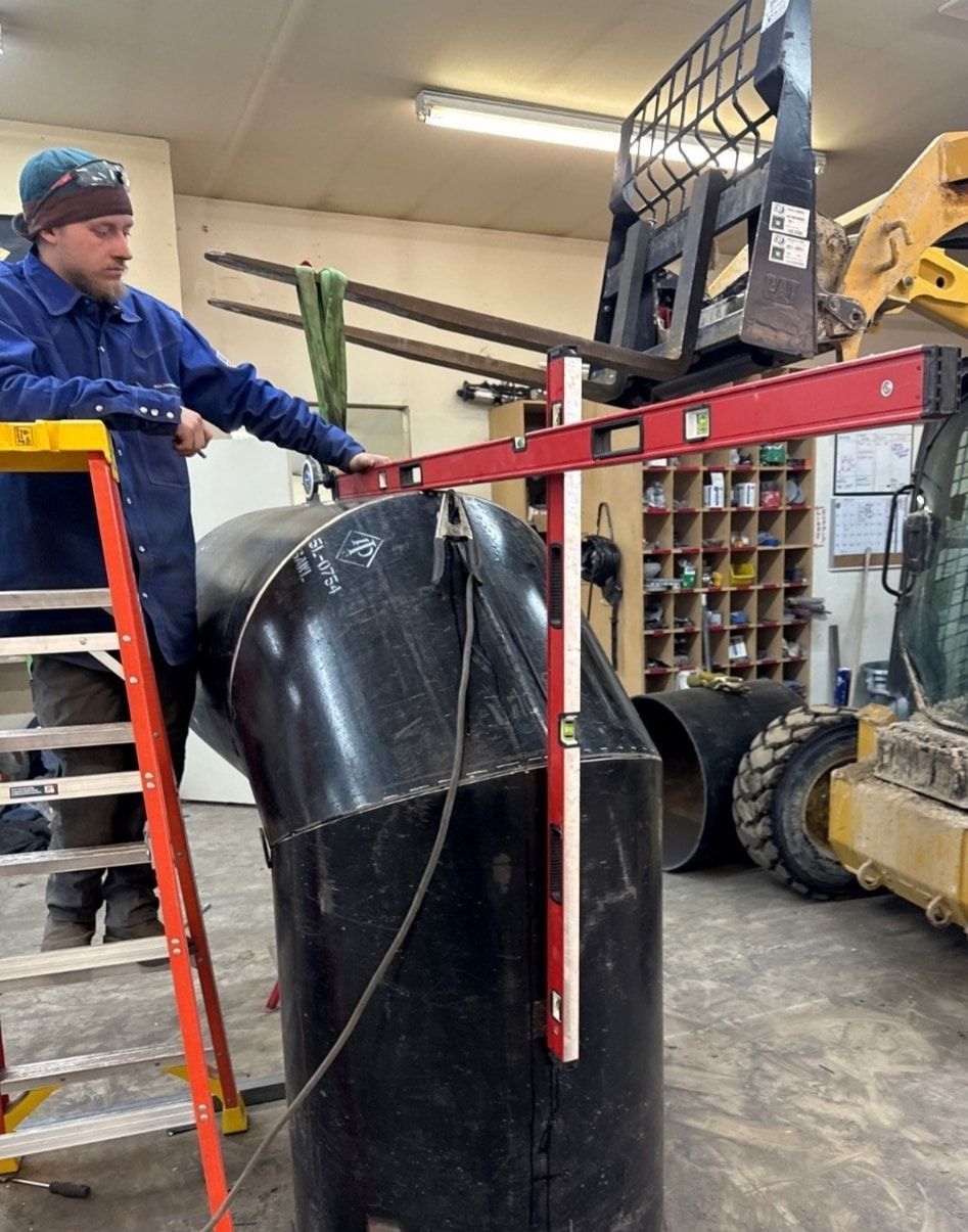 Man on ladder uses level on large pipe in a workshop. Forklift nearby.