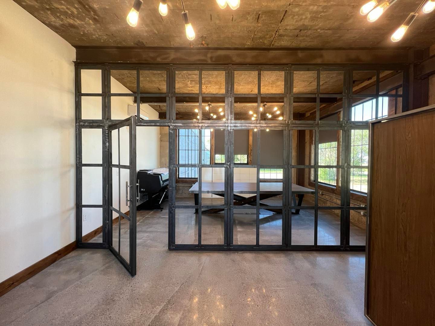 Industrial-style office space with glass-paned room dividers.  Concrete floor, exposed brick, and a meeting table are visible.