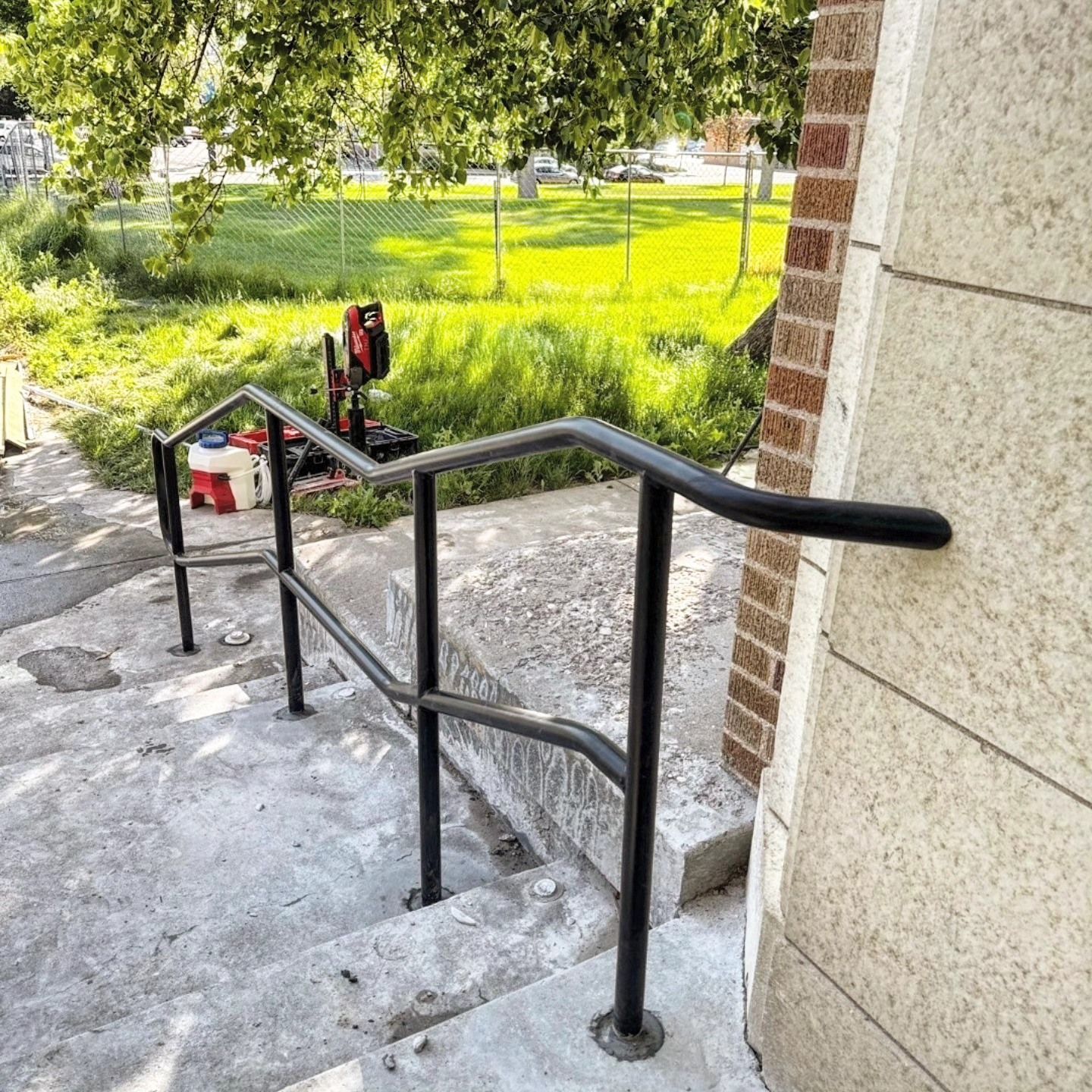 Black handrail on concrete steps next to a building, with grassy area in the background.