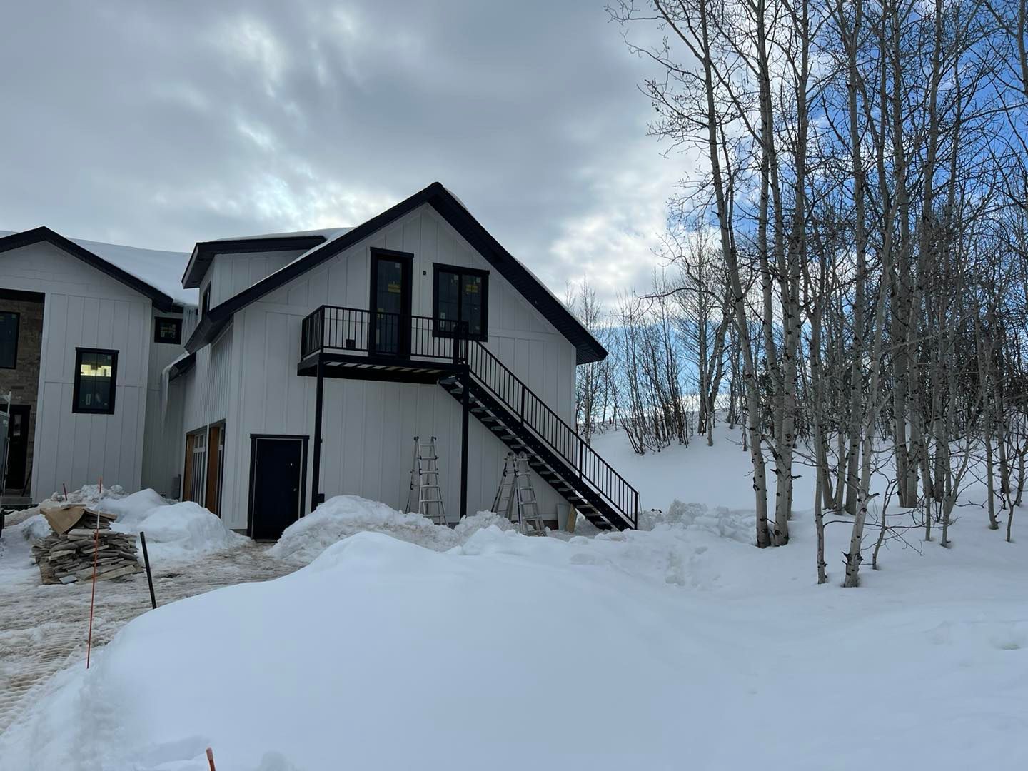 White house with black trim and outdoor staircase, covered in snow, under cloudy sky.