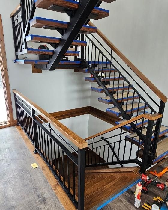 Staircase with wooden steps and railing, black metal balusters. Construction in progress, blue tape visible.