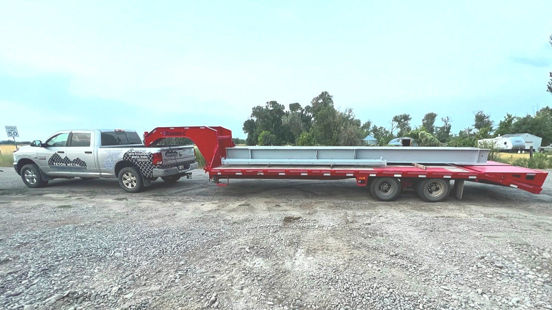 Gray truck towing a red trailer with a long metal beam on a gravel lot under a cloudy sky.