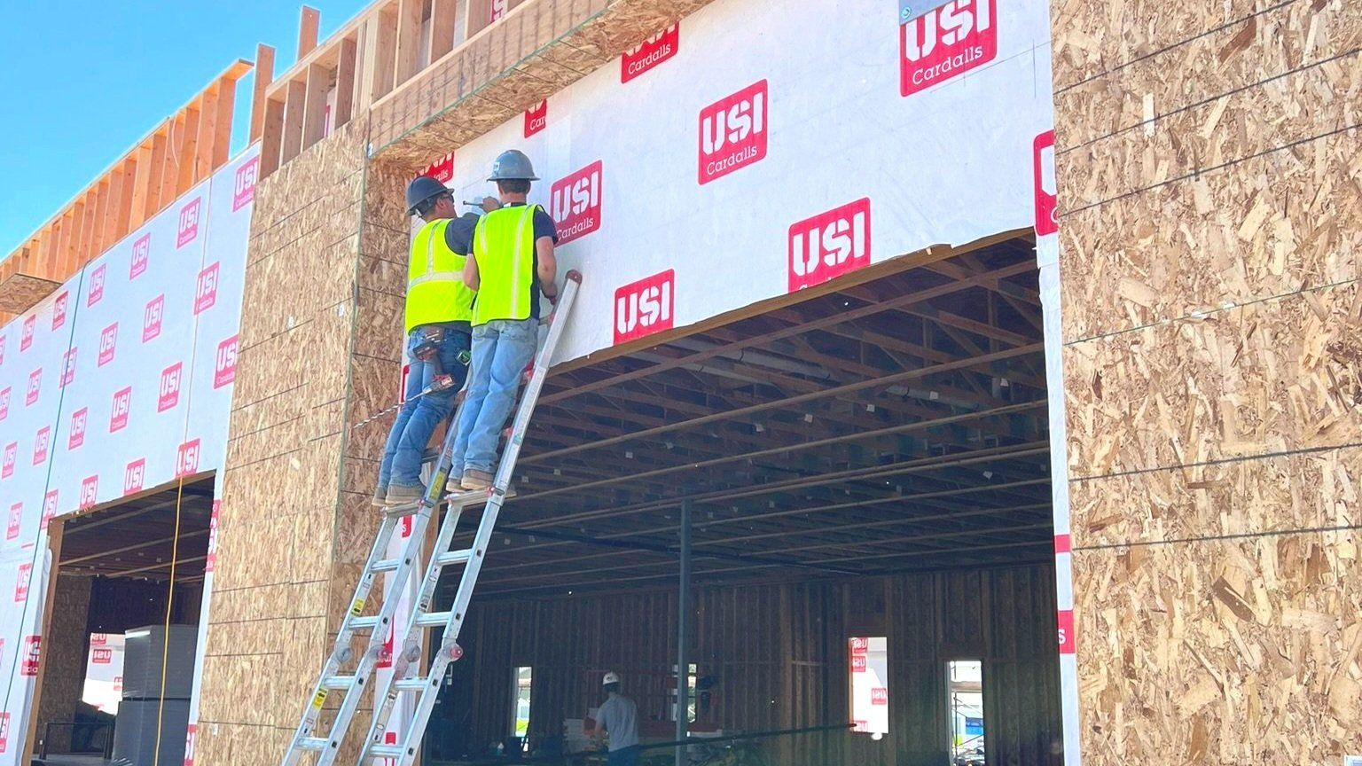 Construction workers installing weather barrier on a building facade; one on a ladder.