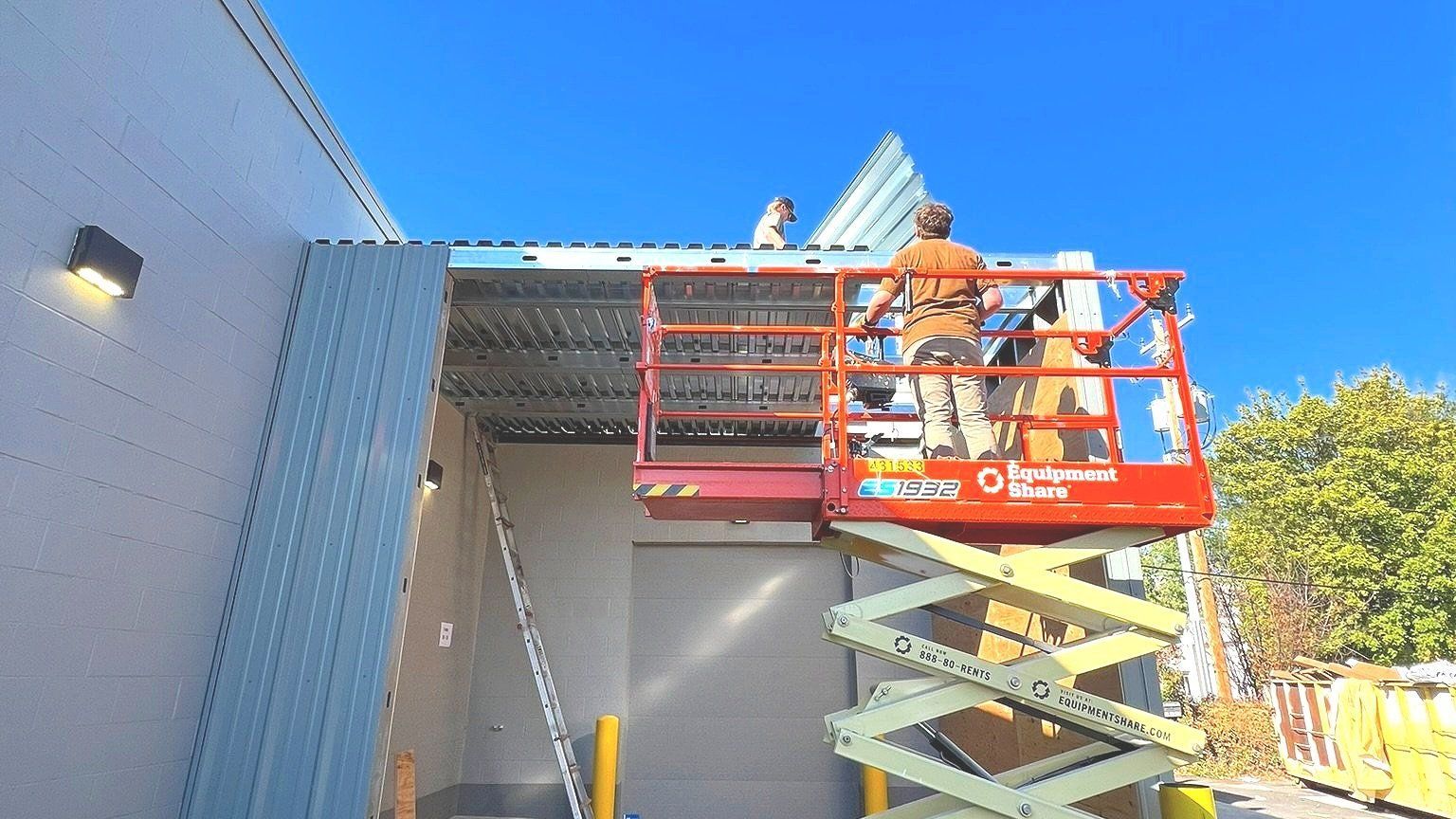 Two workers on a scissor lift install blue corrugated metal siding on a building's exterior under a clear blue sky.
