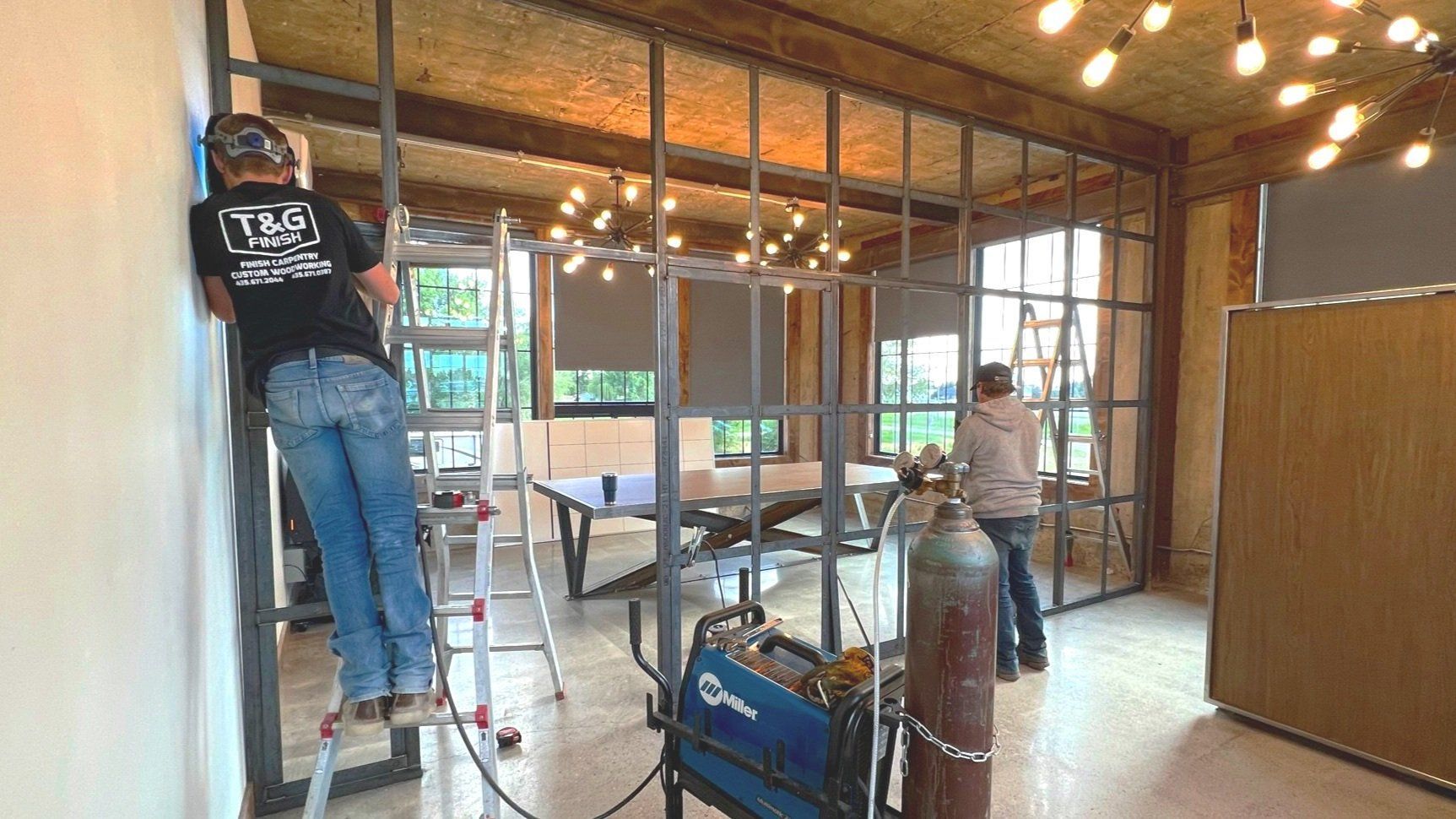 Workers welding a steel-framed interior wall. One is on a ladder; a welding machine and gas tank are nearby.