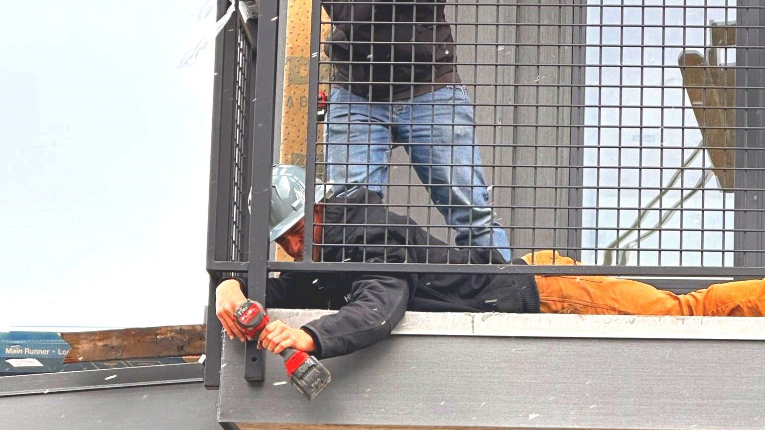 Construction worker using a drill while lying in a caged area. Another worker stands above.