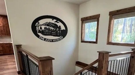 Metal sign with cabin design on a white wall in a home stairwell, with windows and a wooden railing.
