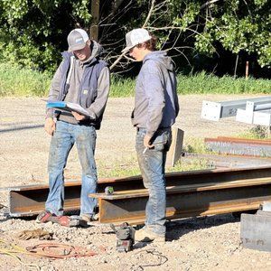 Two workers on a construction site, looking at a clipboard. They are near steel beams and wear work clothes and hats.