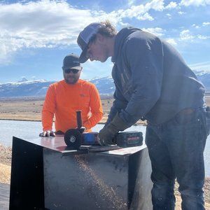 Two men working outdoors, one using a grinder on a black metal box, blue sky, mountains, and water in the background.
