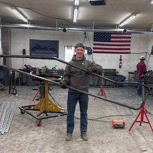 Man in a workshop holding a long, bent metal frame. American flag and welding equipment in the background.