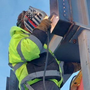 Welder in neon jacket welding metal beams outdoors. Welding helmet with flag design.