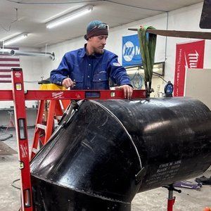 Man in blue work clothes using a level on a large black pipe in a workshop.