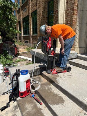 Man in orange shirt drills concrete steps outside a building. Drill, water tank, and tools are present.