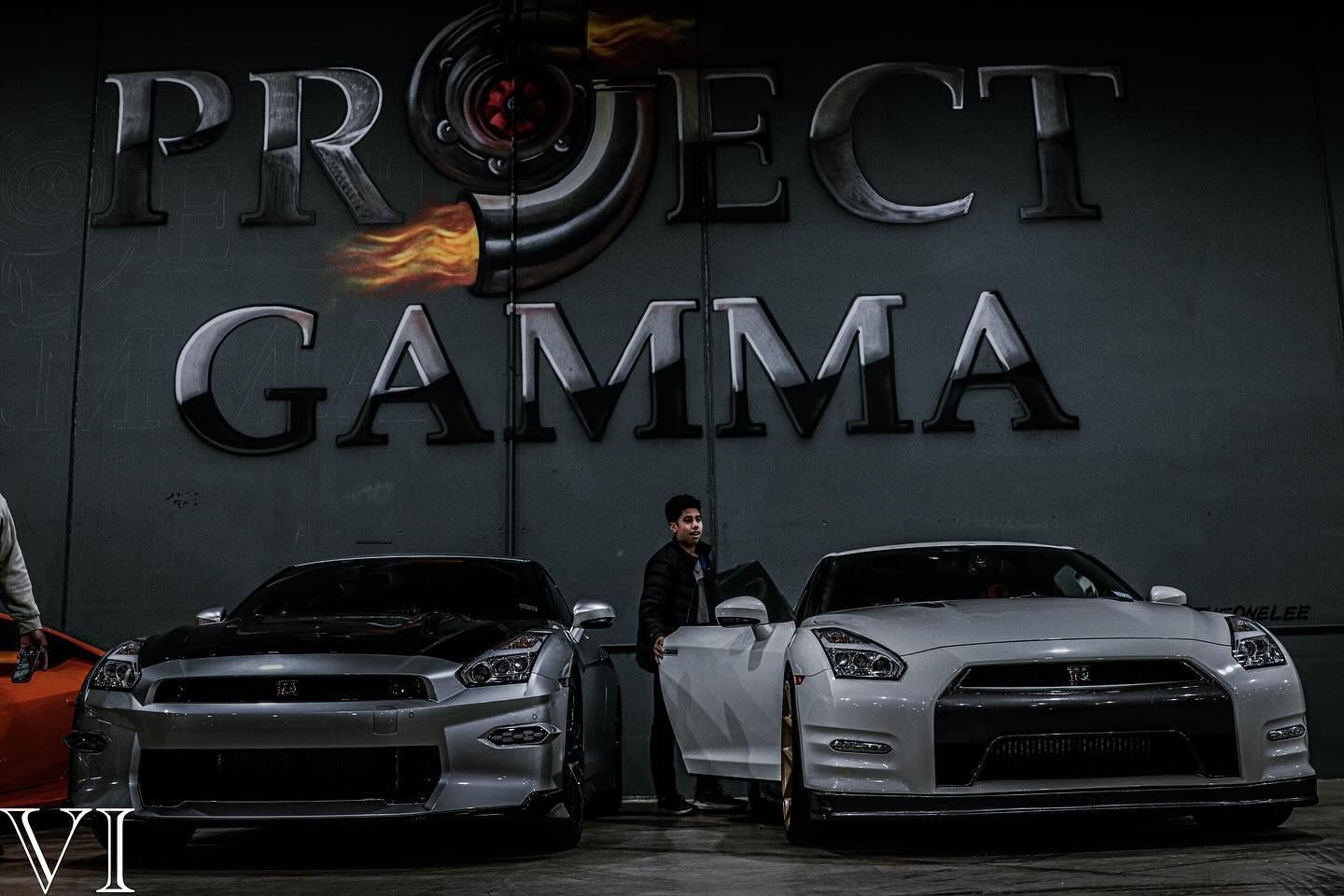 A man is standing next to two sports cars in front of a project gamma sign.