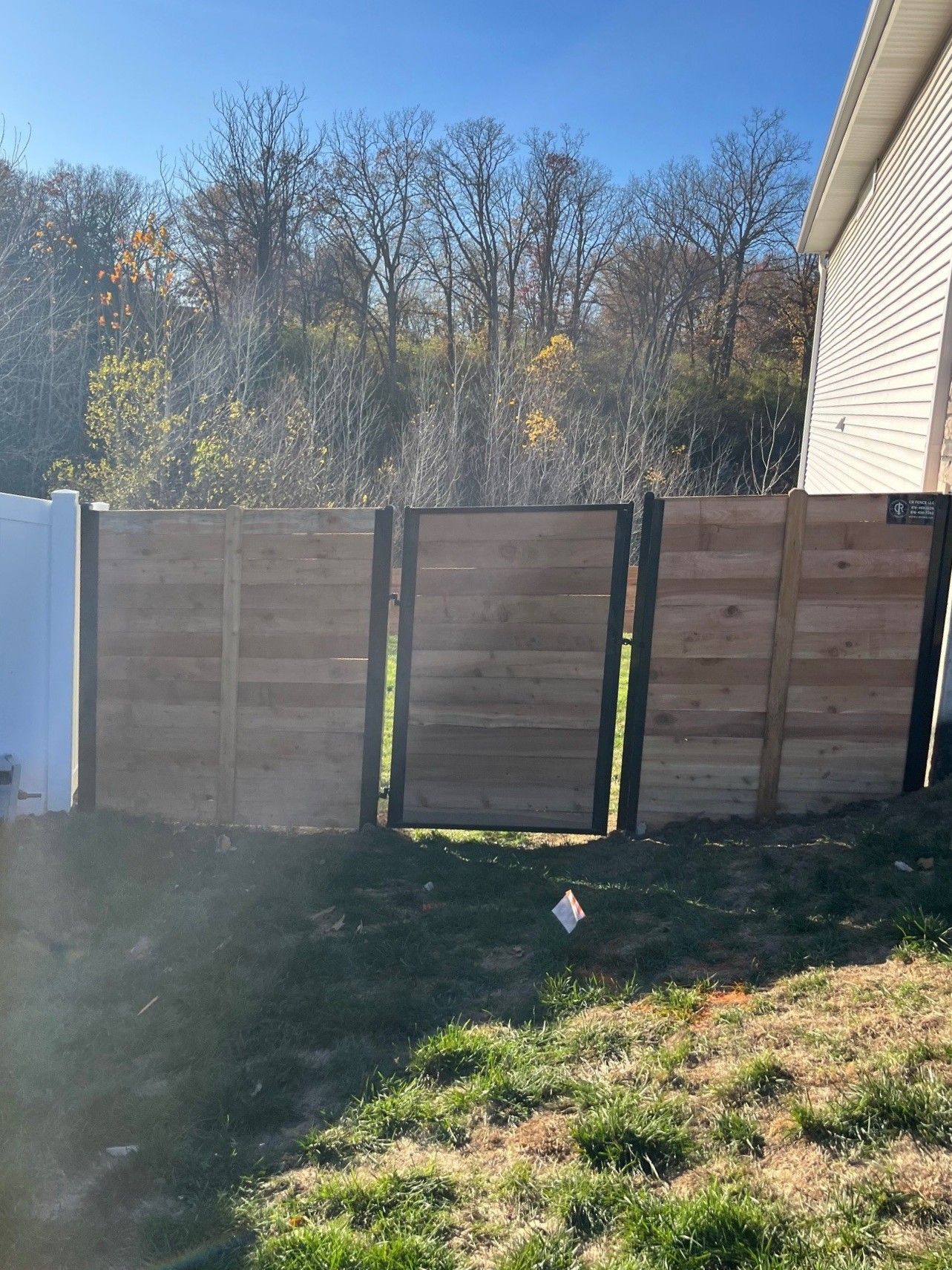A wooden fence gate with black metal framing stands on a grassy slope next to a white fence and the side of a house.