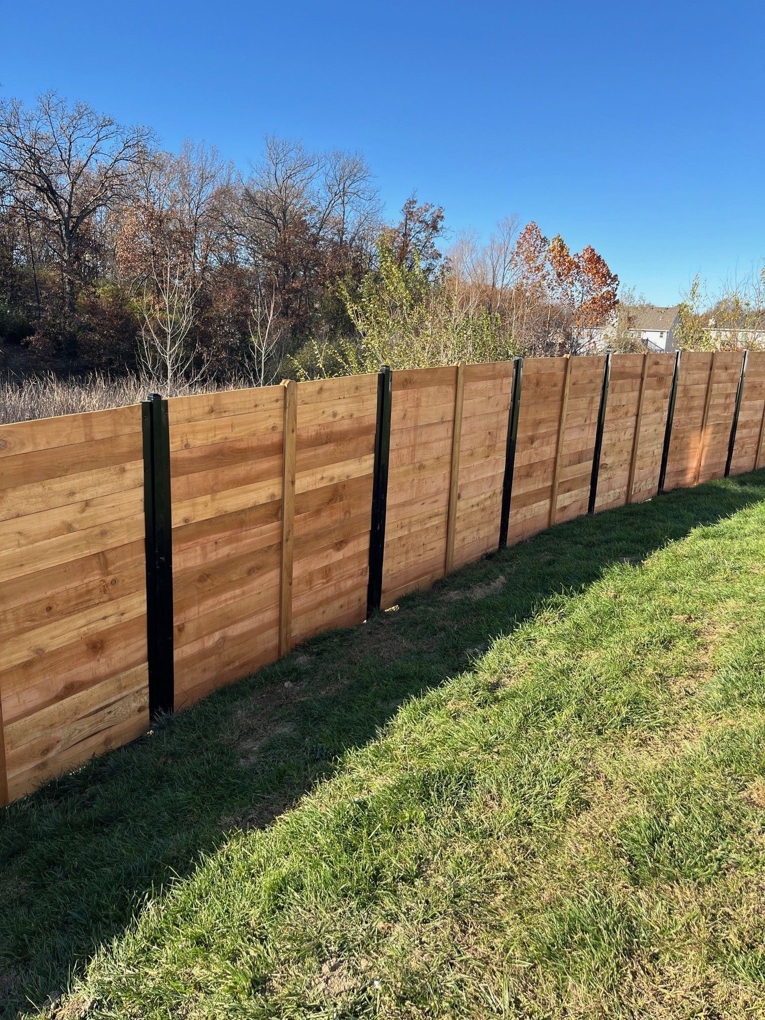 A horizontal wooden fence with black posts stretches across a grassy lawn under a clear blue sky.
