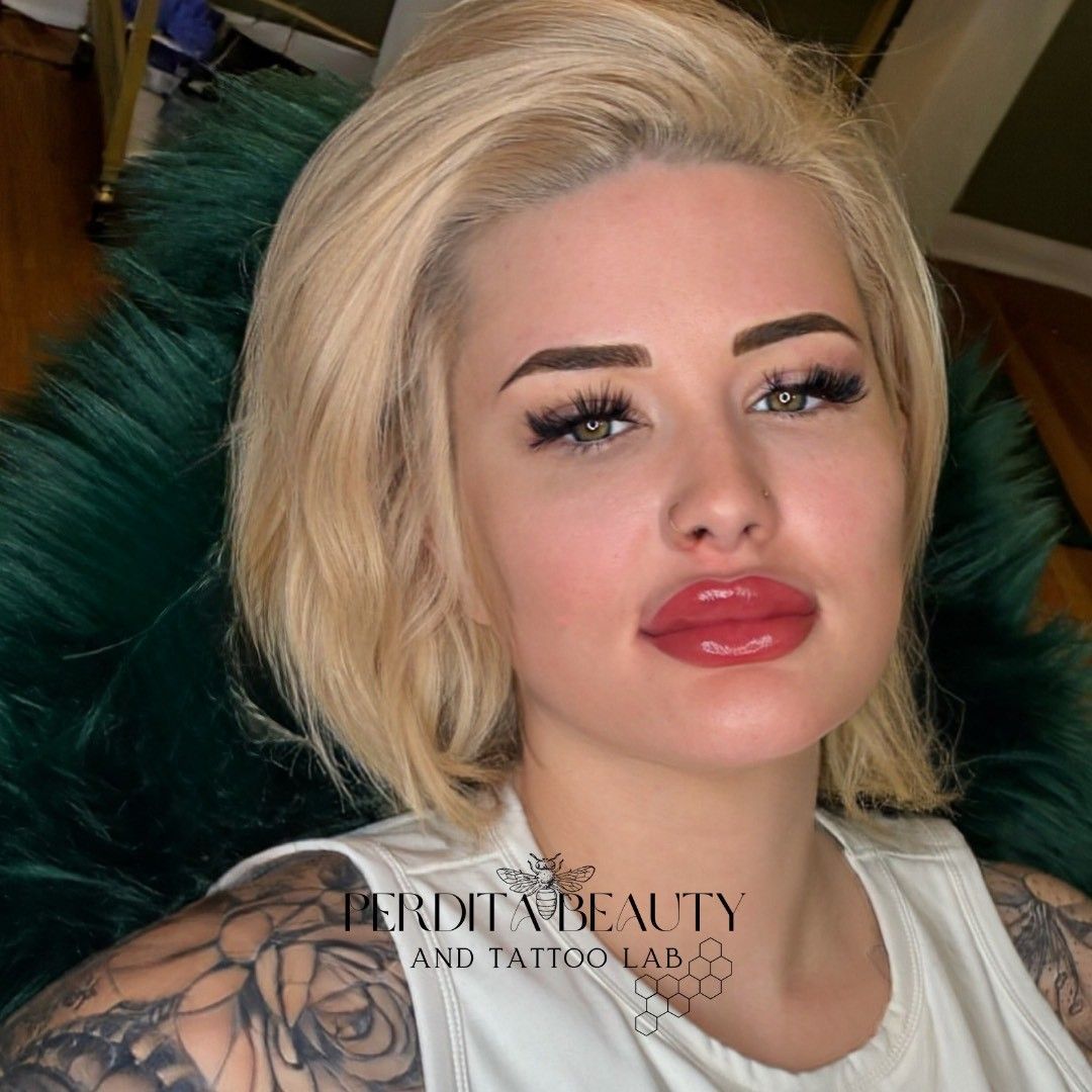 Blonde woman with full lips, dark eyebrows, and arm tattoos, seated; 