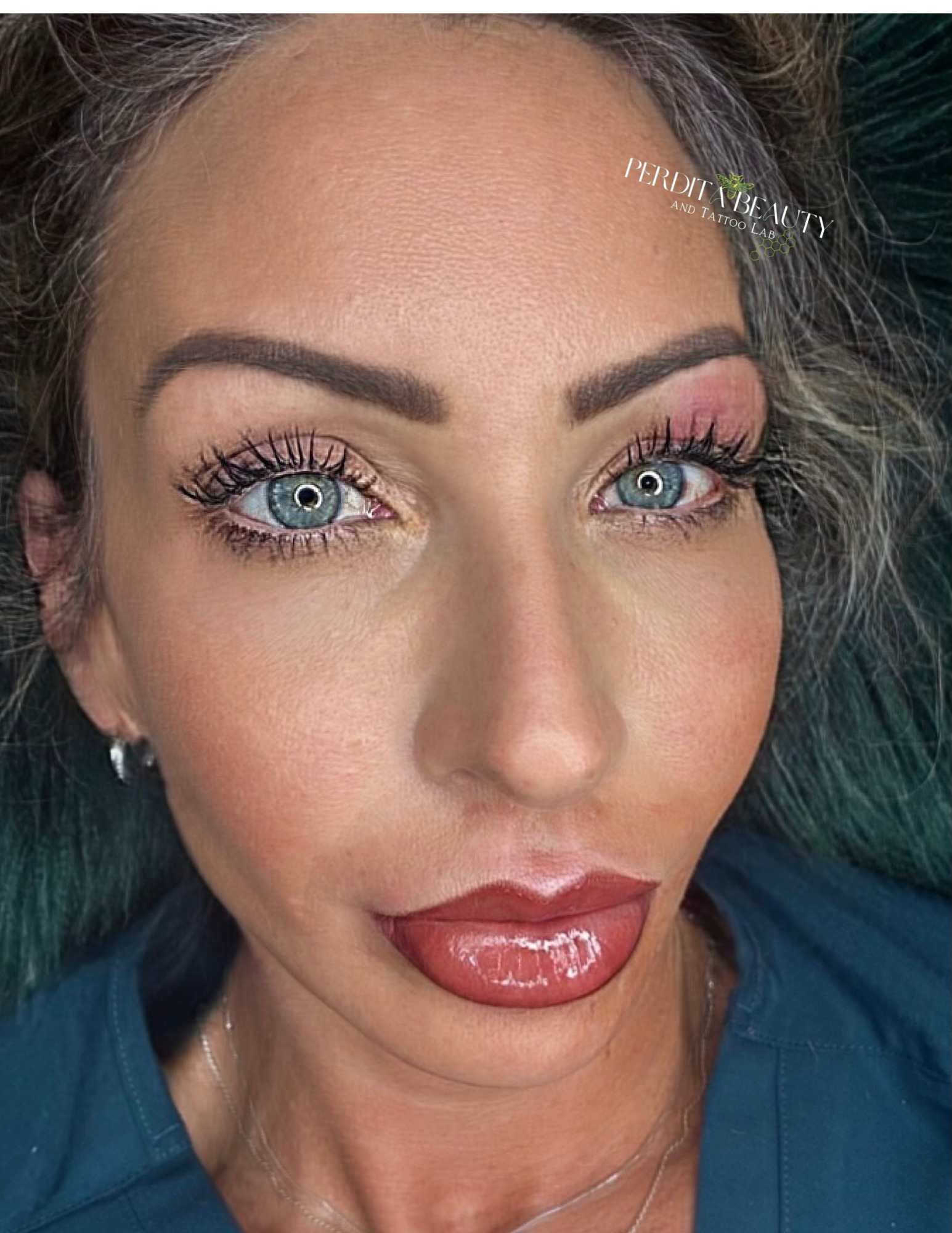 Close-up of a person with blue eyes, dark hair, pink eye makeup, and glossy lips against a dark background