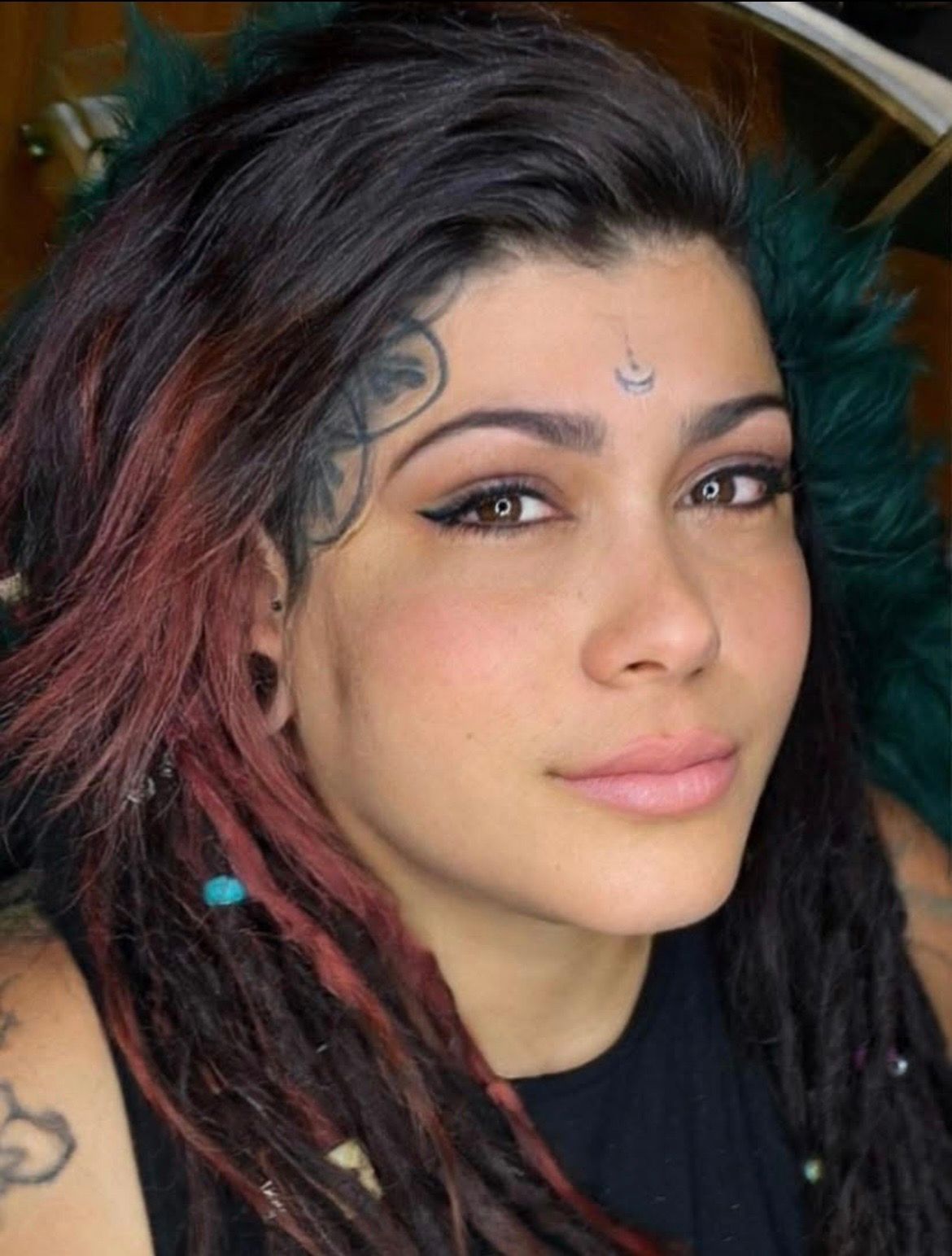 Close-up portrait of a person with dark hair, reddish streaks, and small facial tattoos, looking at the camera.