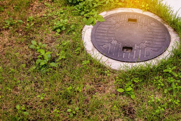 Round manhole cover in grass beside a paved edge, with sunlight and moss around it