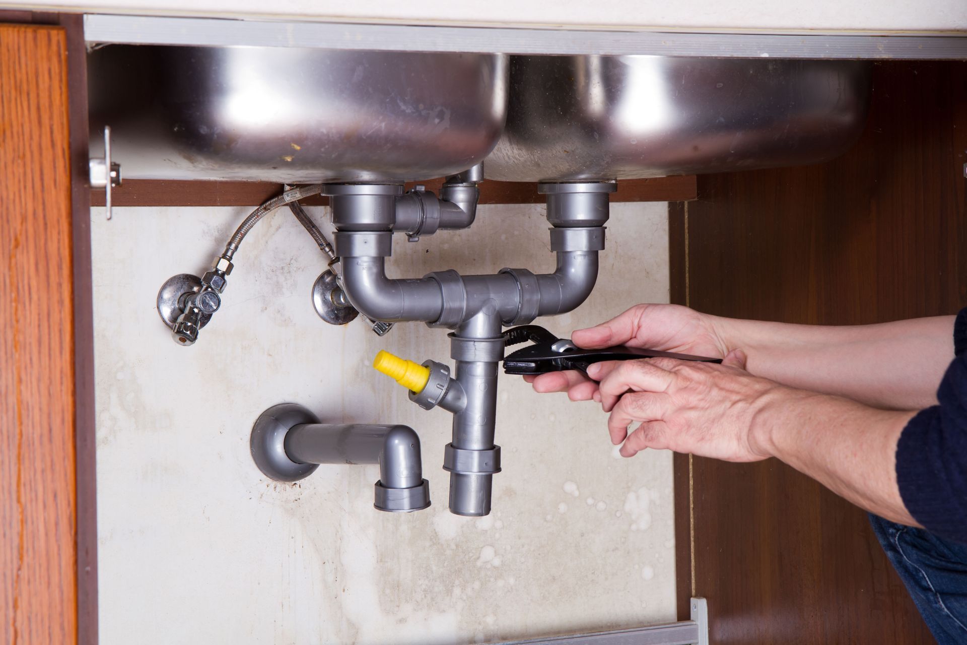 Hands using a wrench on plumbing under a kitchen sink. Silver pipes, yellow valve, and metal basins are visible.