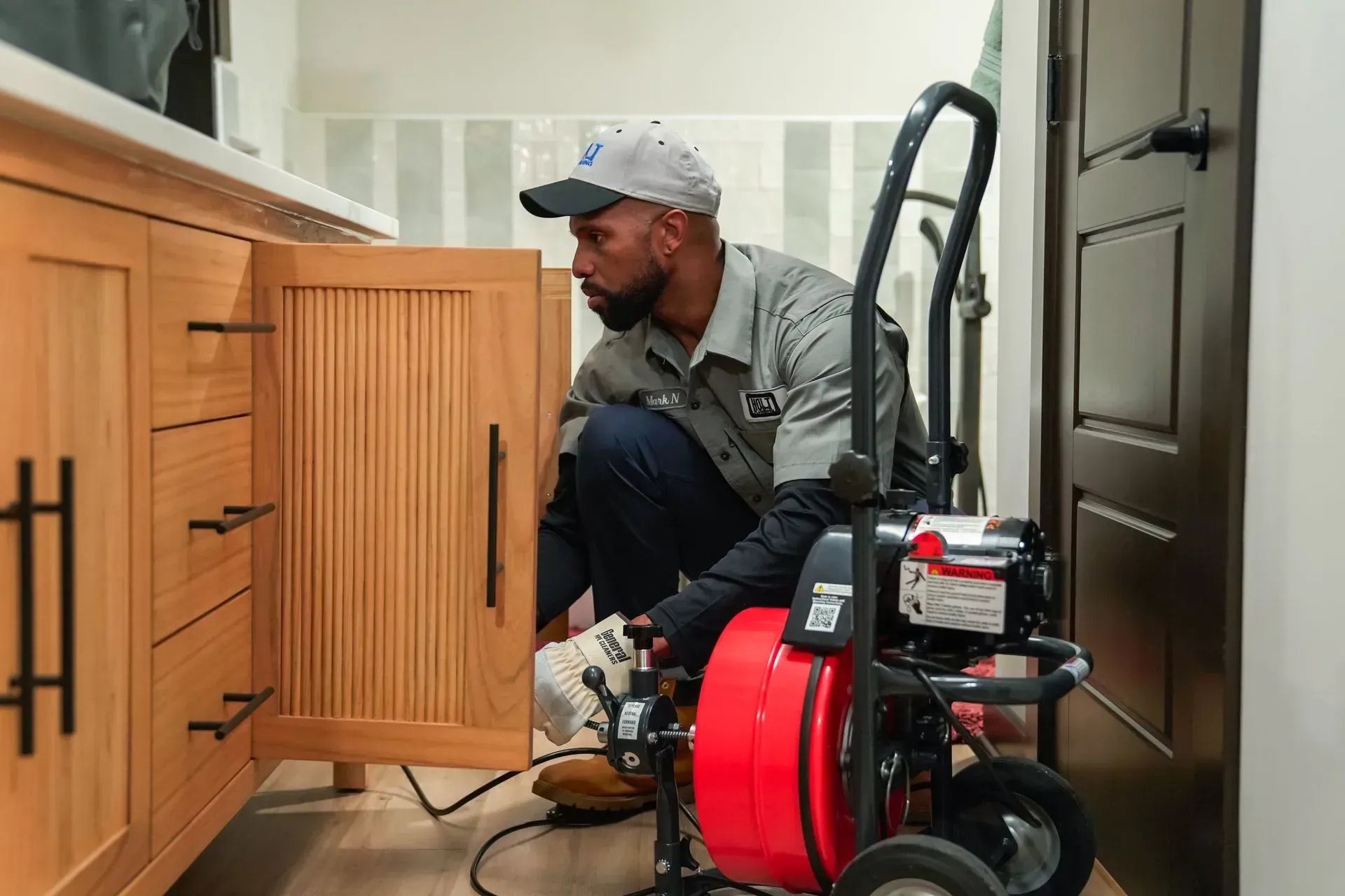 Plumber working on a sink, using a drain snake in a bathroom with wooden cabinets.