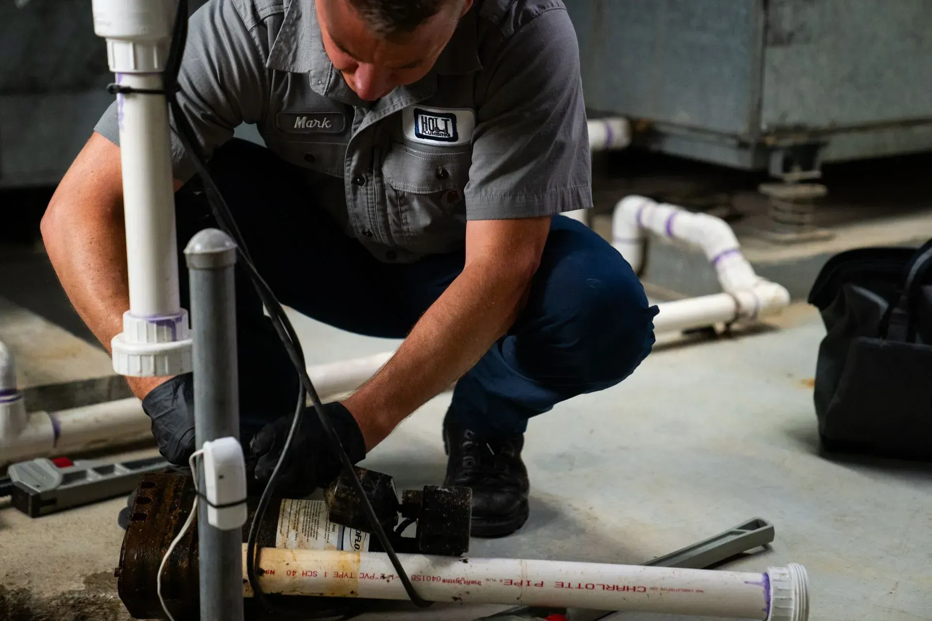 Plumber in gray uniform, crouched, working on PVC pipes in a basement, black gloves.