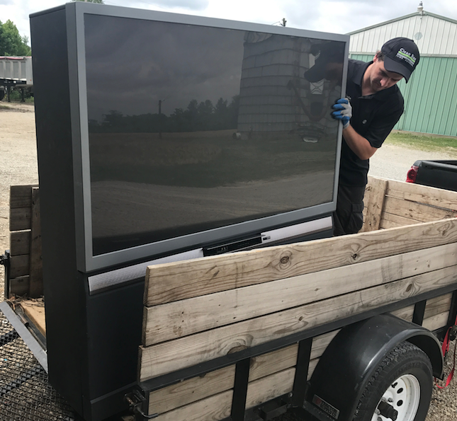 A man loads a large TV into a wooden trailer in a rural setting.