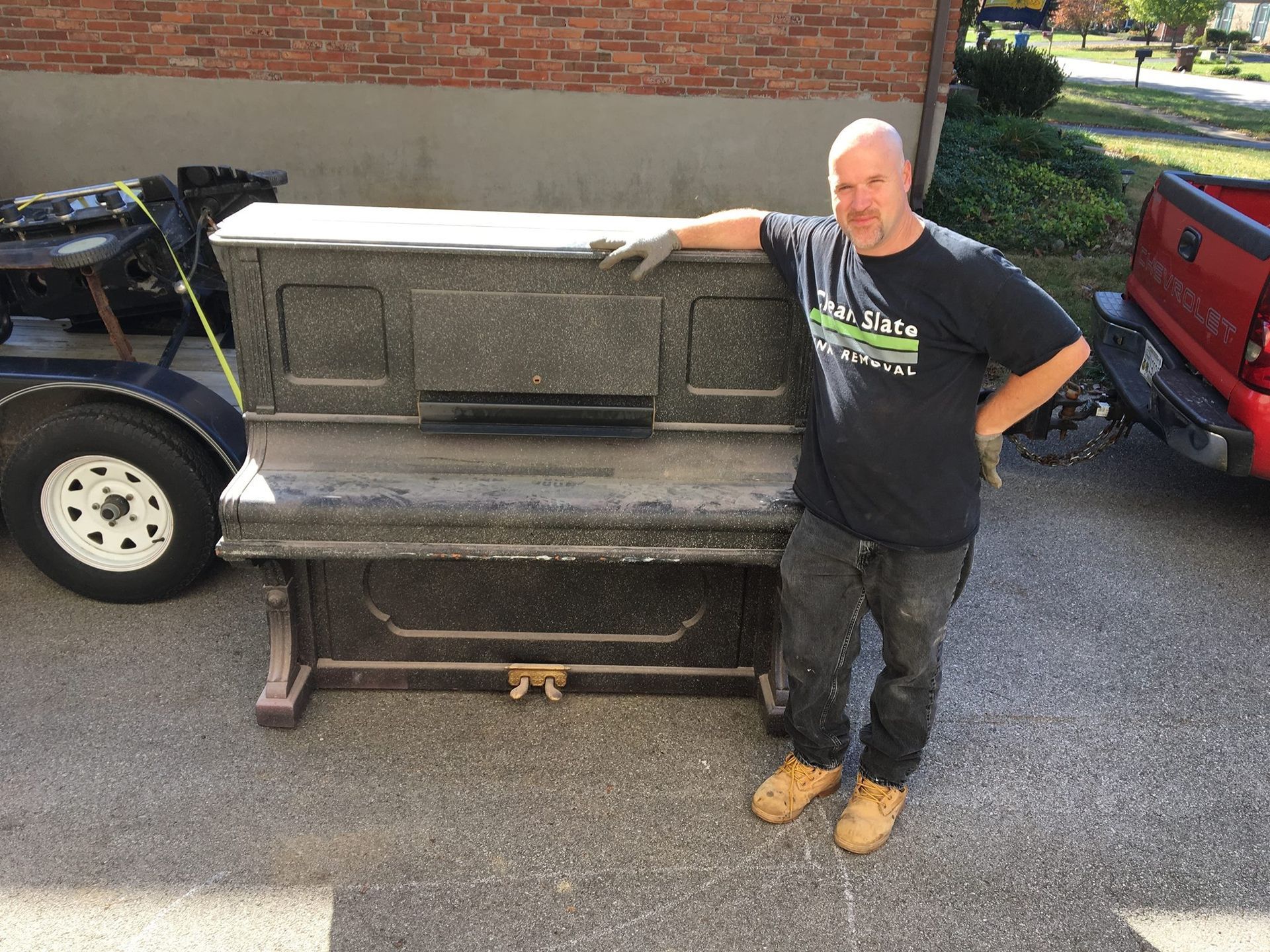 Man standing next to a weathered, dark piano on a driveway, next to a truck and trailer.
