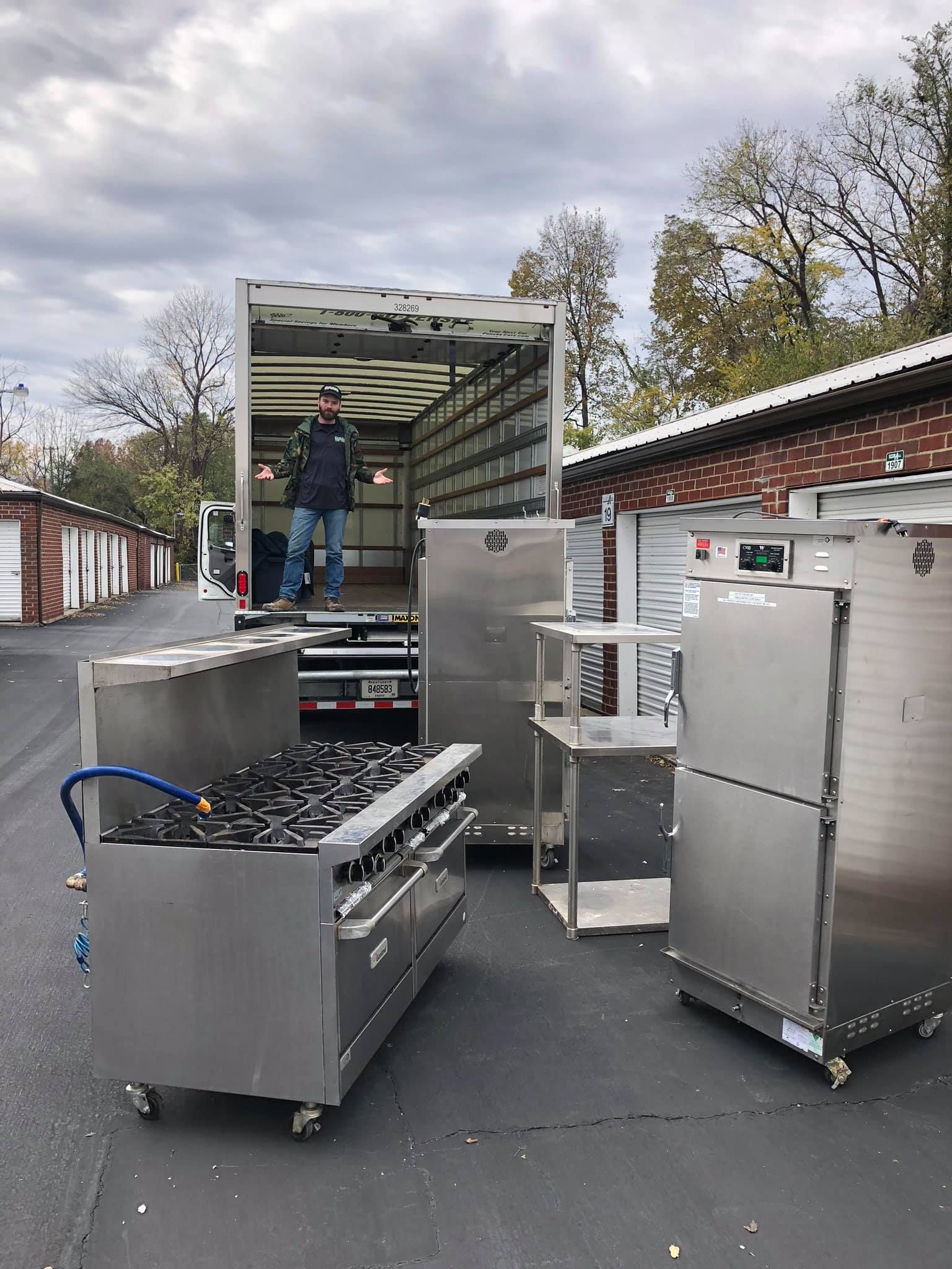 Man unloading commercial kitchen equipment from a truck in an alley; stainless steel appliances.
