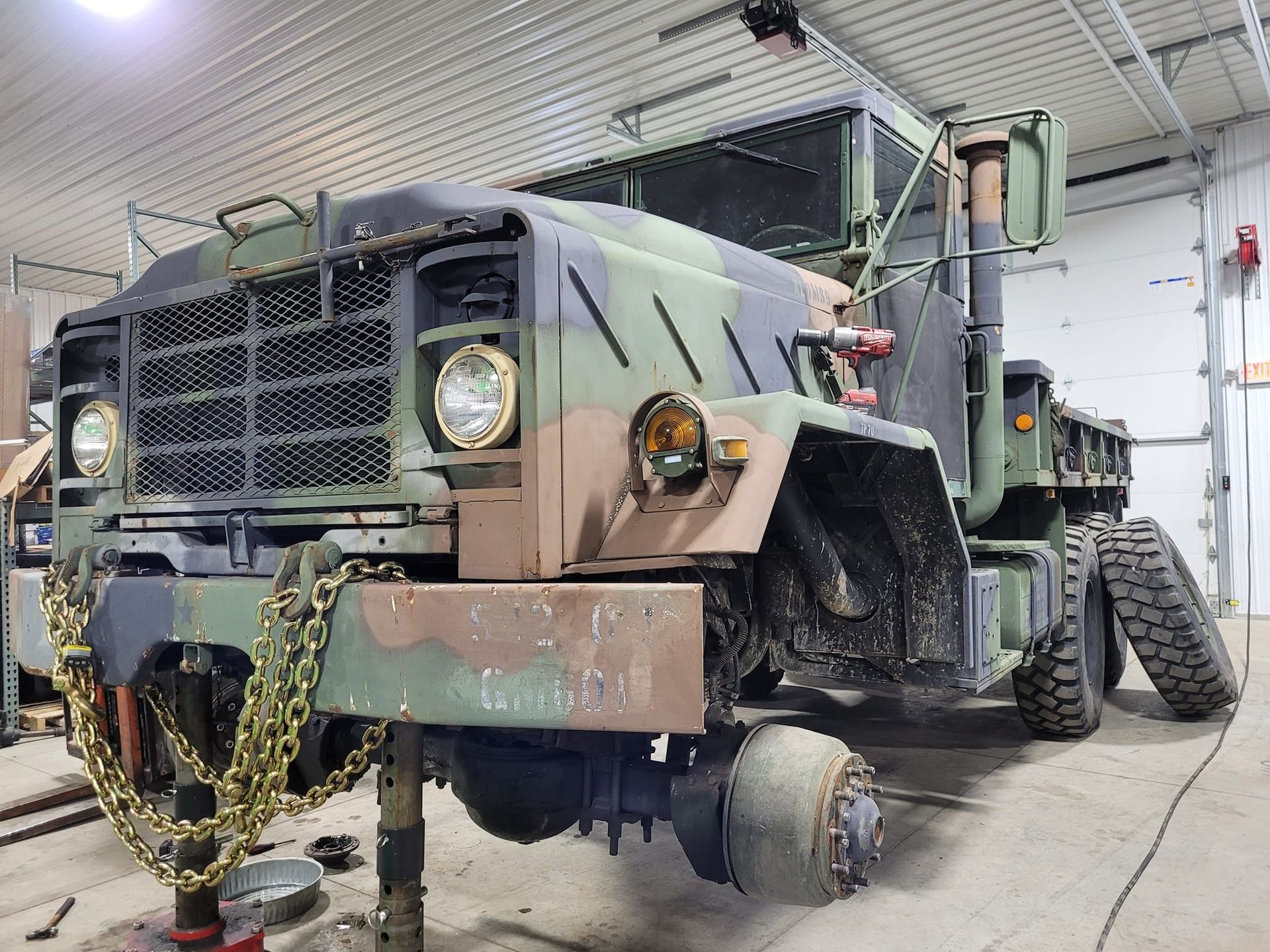 Camouflaged military truck being worked on in a garage. Front end raised, no tires.