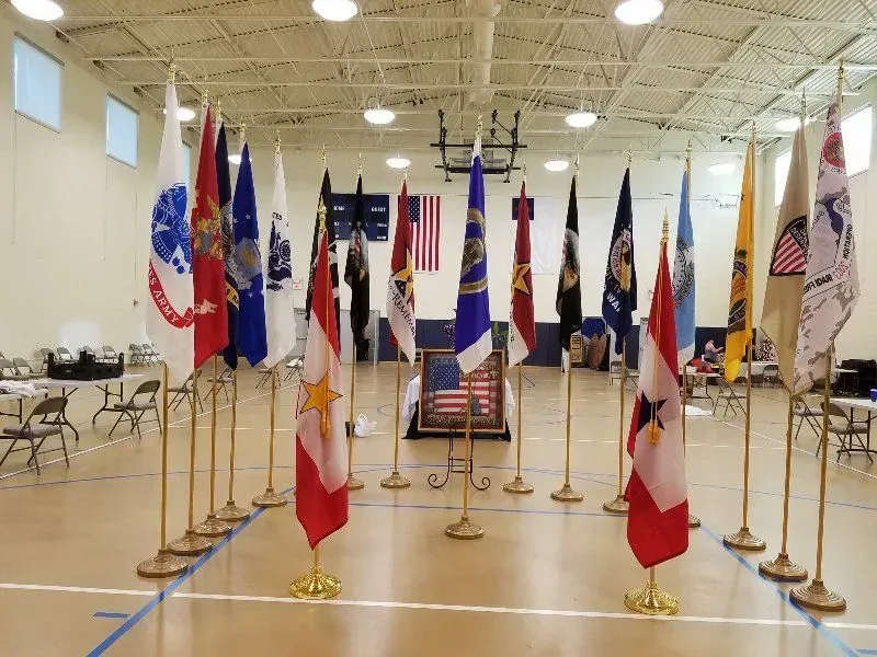 Flags on display in a gymnasium. Multiple military and US flags stand in a semi-circle.