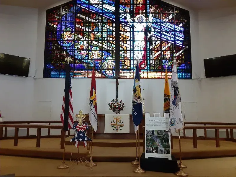 Altar with flags, stained glass window, and cross in a church. American flag and others stand in front.