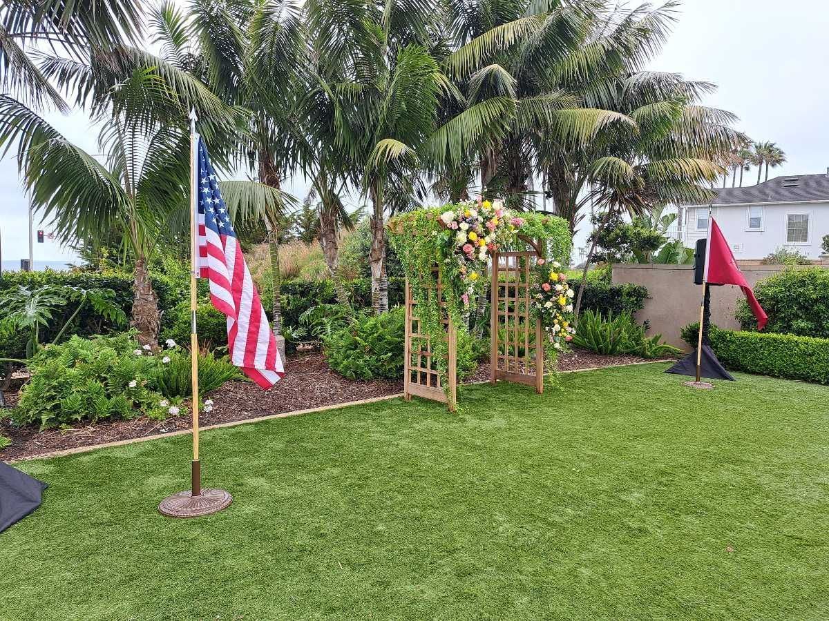 American flag, floral arch, and greenery on a lawn, possibly for a ceremony, with palm trees in the background.