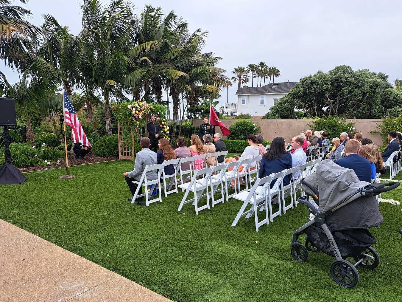 Outdoor wedding ceremony with guests seated in white chairs on a lawn. An American flag stands near the ceremony.