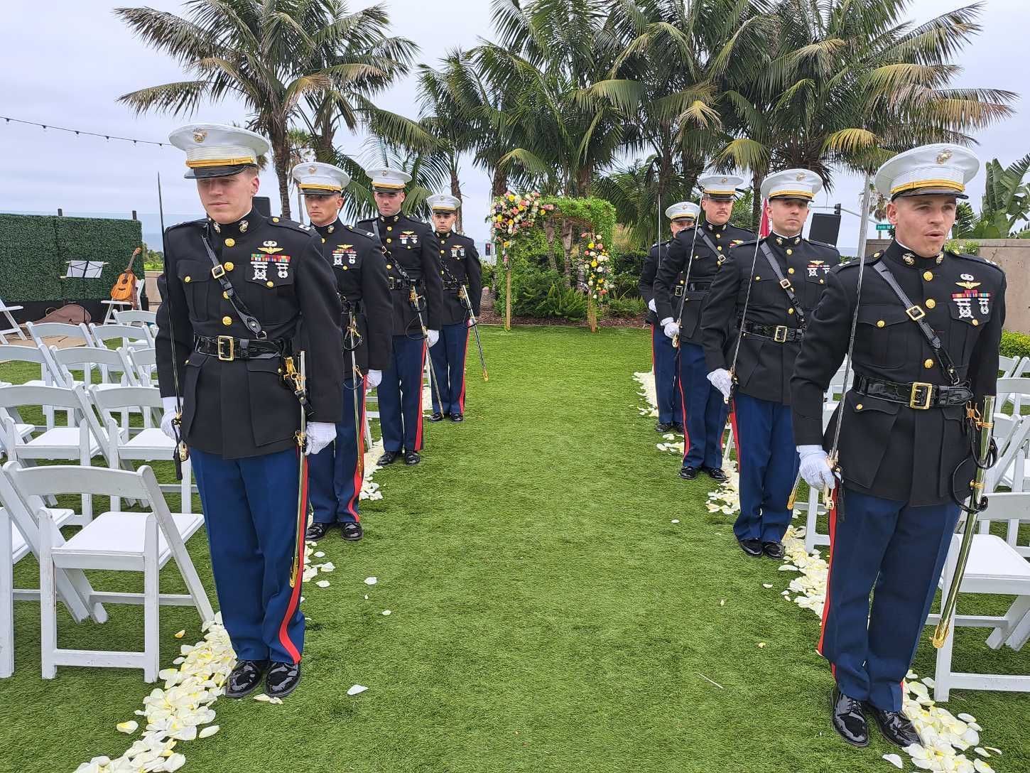 Marines in dress blues stand in formation on grass pathway at a ceremony, white chairs on the sides.