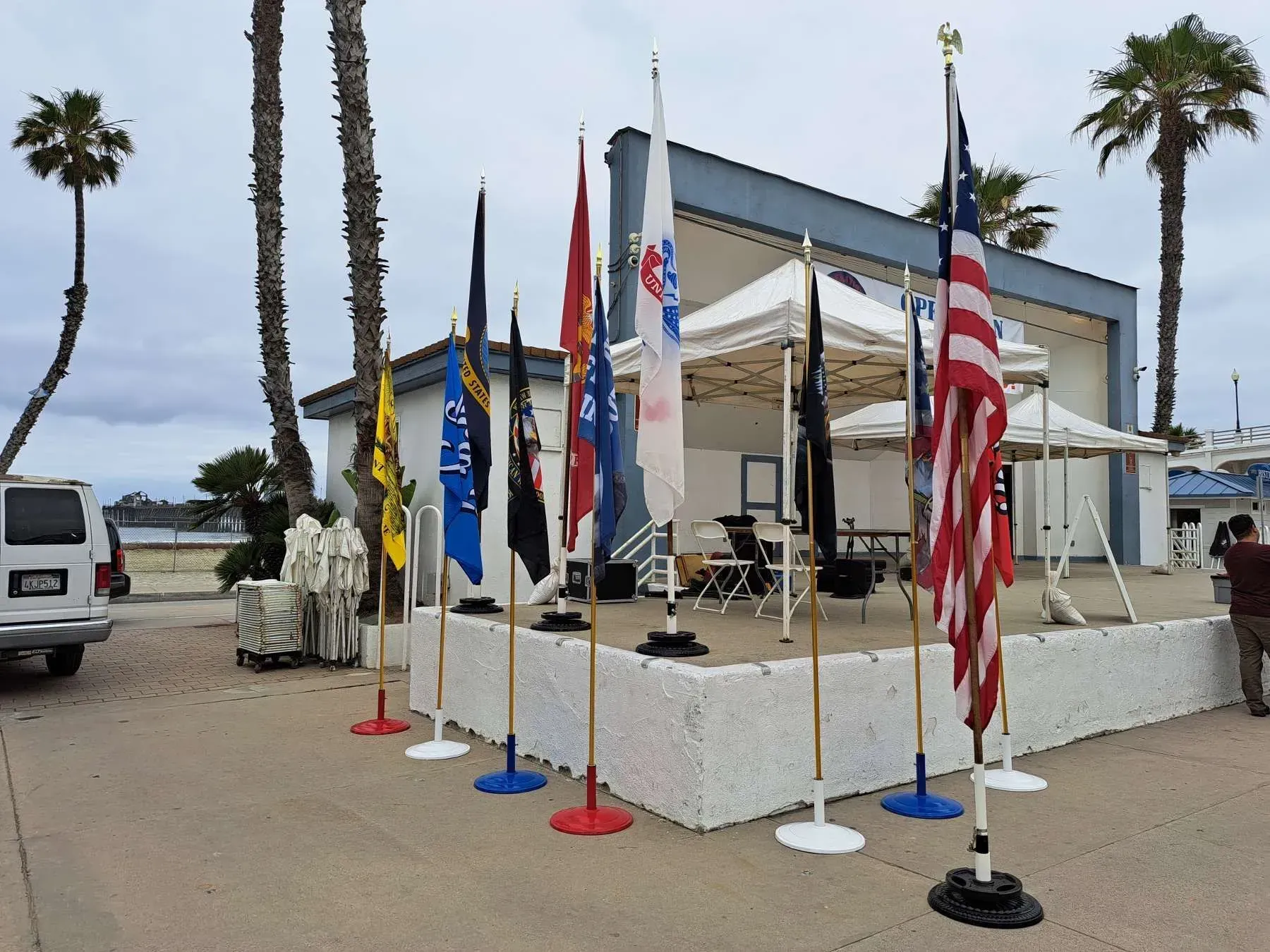 Flags on display in front of a blue building near the coast, under a cloudy sky.